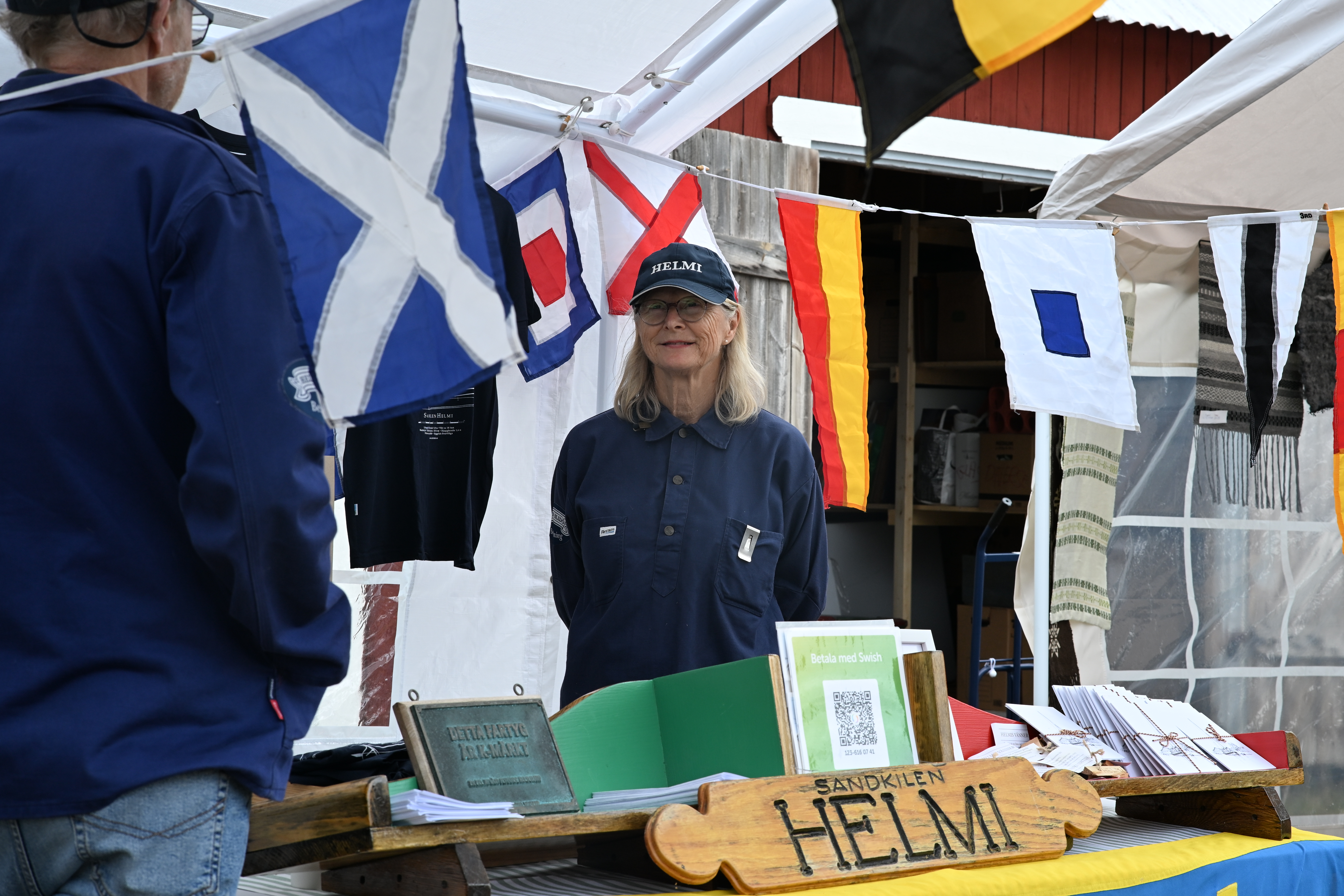 En kvinna i seglar skjorta och keps med texten Helmi står bakom ett marknadsbord.