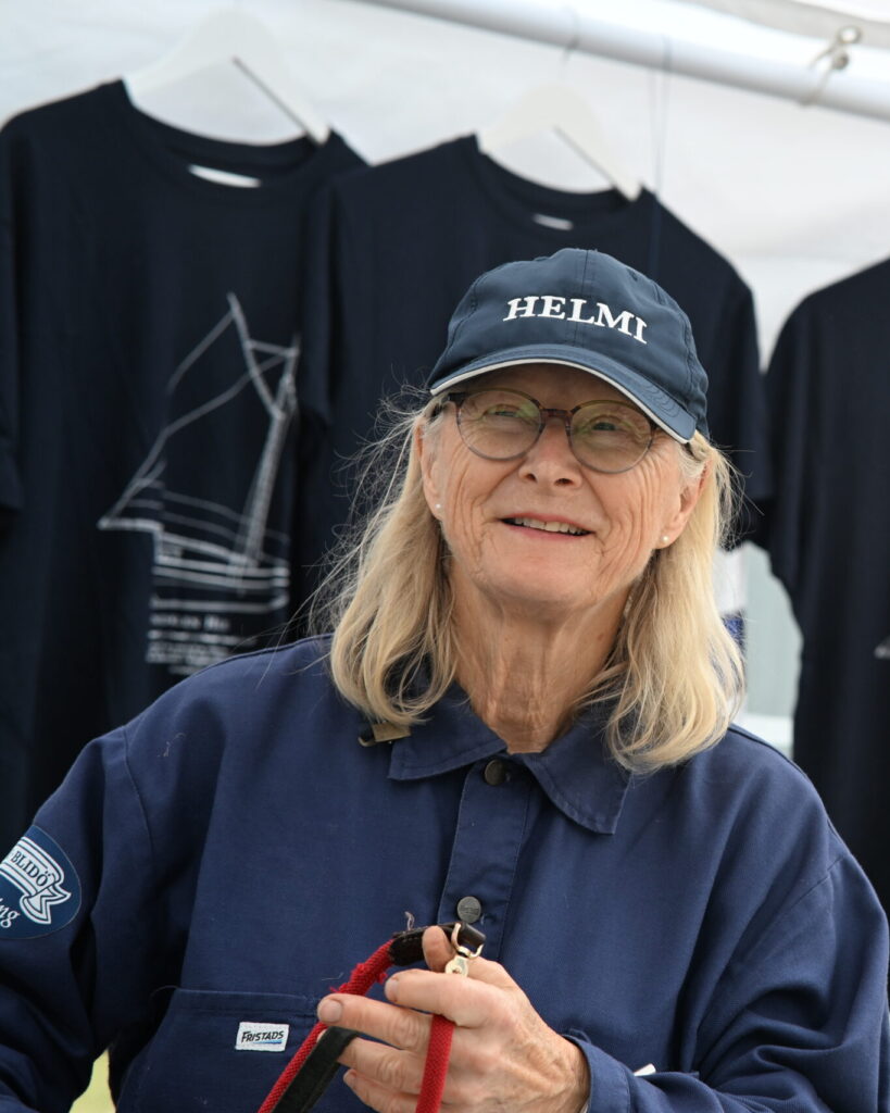 En kvinna i seglarskjorta och keps, med texten Helmi. Hon håller i en röd tamp och bakom henne hänger t-shirts med skutor på.