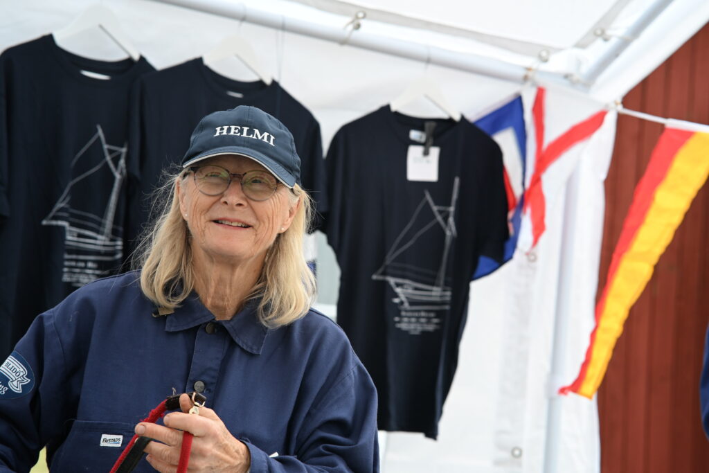 En kvinna i seglarskjorta och keps, med texten Helmi. Hon håller i en röd tamp och bakom henne hänger t-shirts med skutor på.